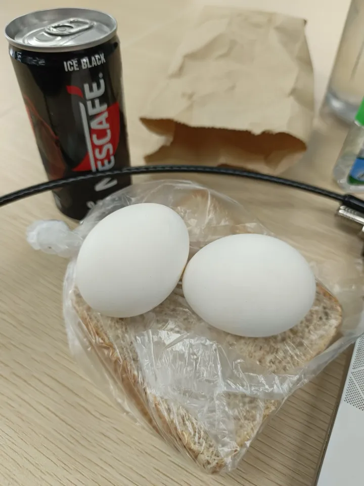 Two boiled eggs on slices of wheat bread in a plastic bag, beside a can of iced black coffee, on an office desk.