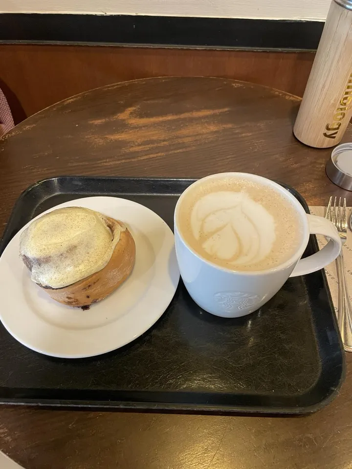A cinnamon roll with icing on a white plate and a latte with leaf art, on a tray on a café table.
